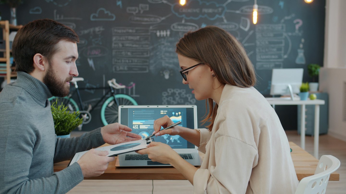 Professionals werken samen aan een laptop en grafieken aan een houten bureau.